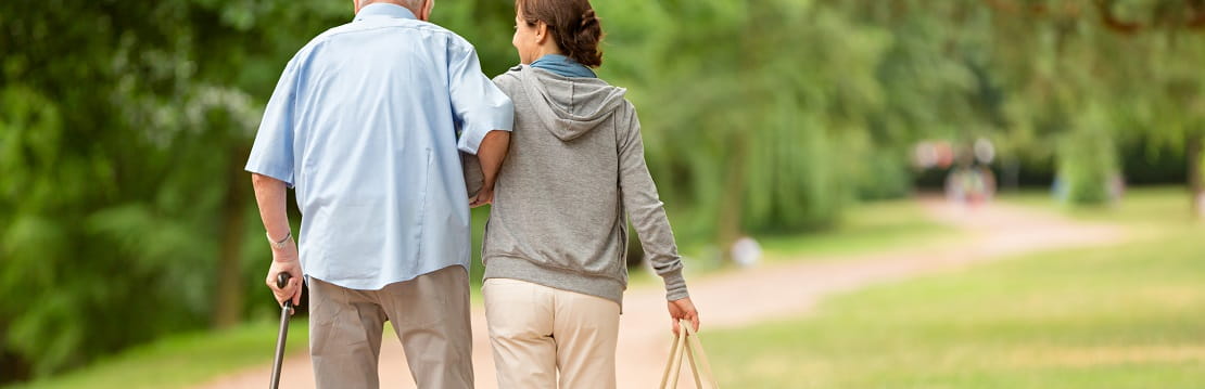 Elderly man and younger woman walk together through a park. 